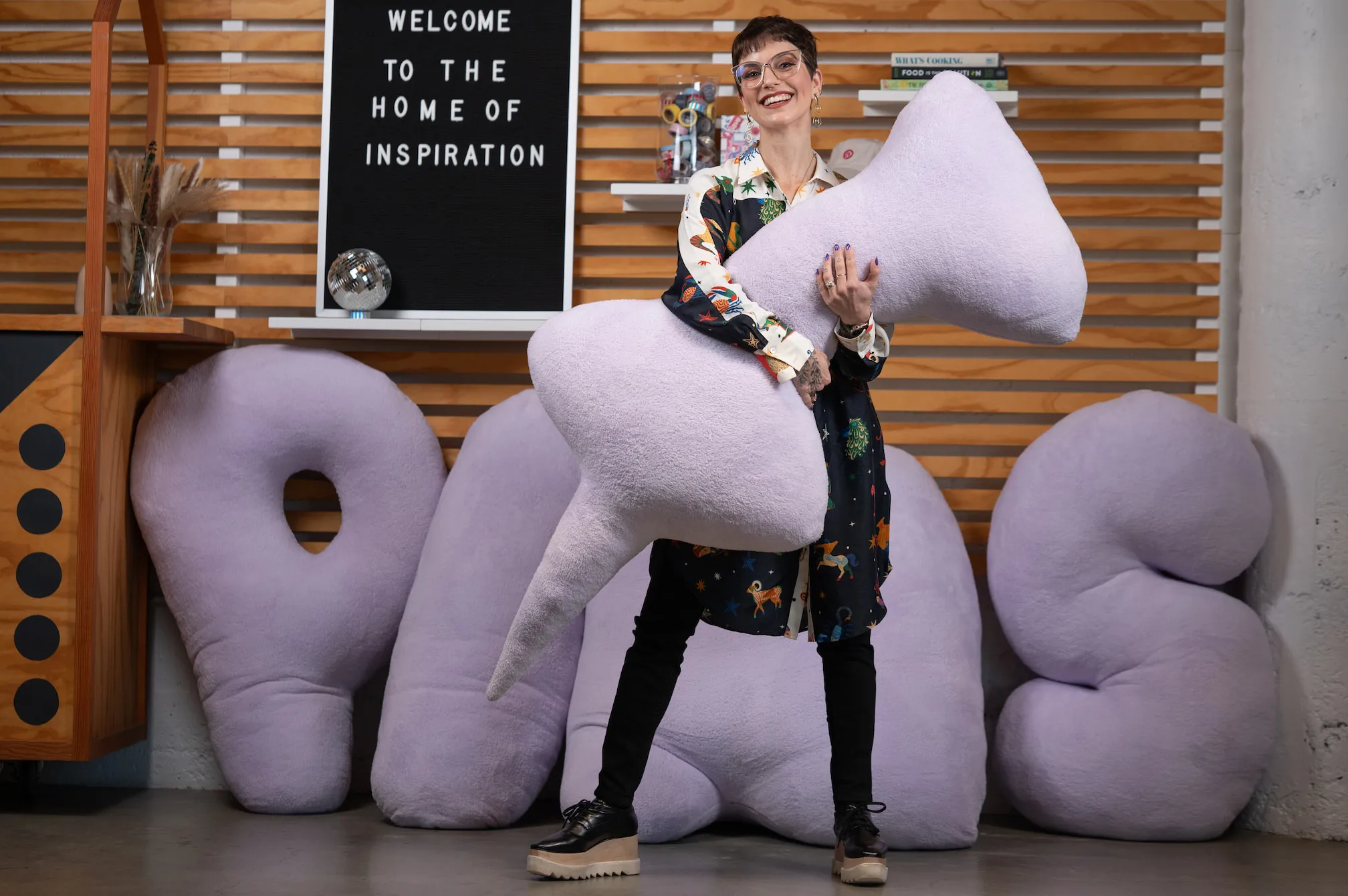 Cintia is standing at the Pinterest office, holding a lilac pin-shaped pillow. She is smiling and surrounded by other pillows that form the word "Pins”. 