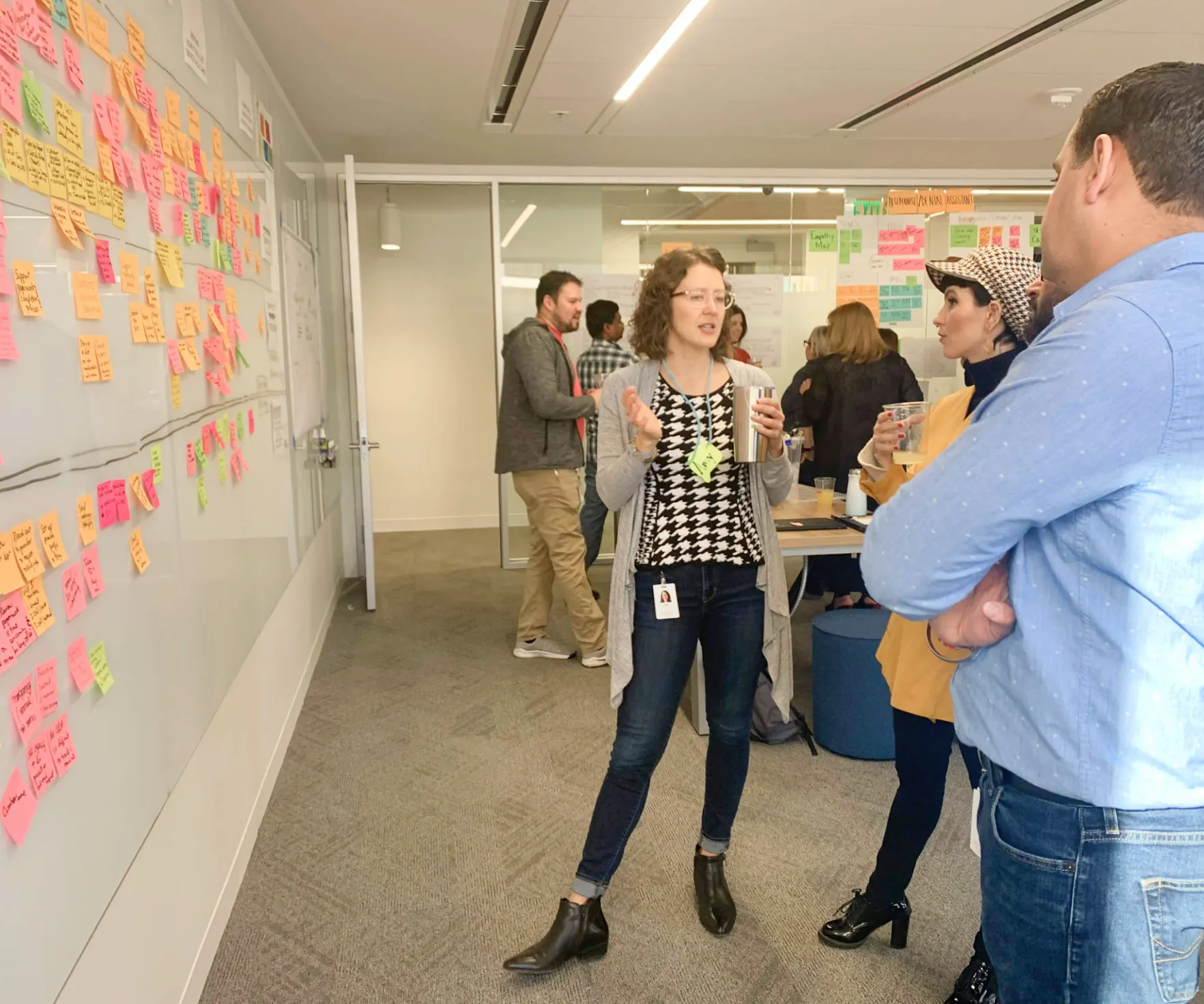 Cintia is standing along 2 other coworkers discussing a journey map. There is a panel full of post-its and notes in front of them. They are at the office attending a workshop event.